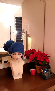 Grand miroir avec une tête à perruque portant un turban bleu avec une fleur cousue dessus et, à côté, un pot de fleur en paille avec un poinsettia 