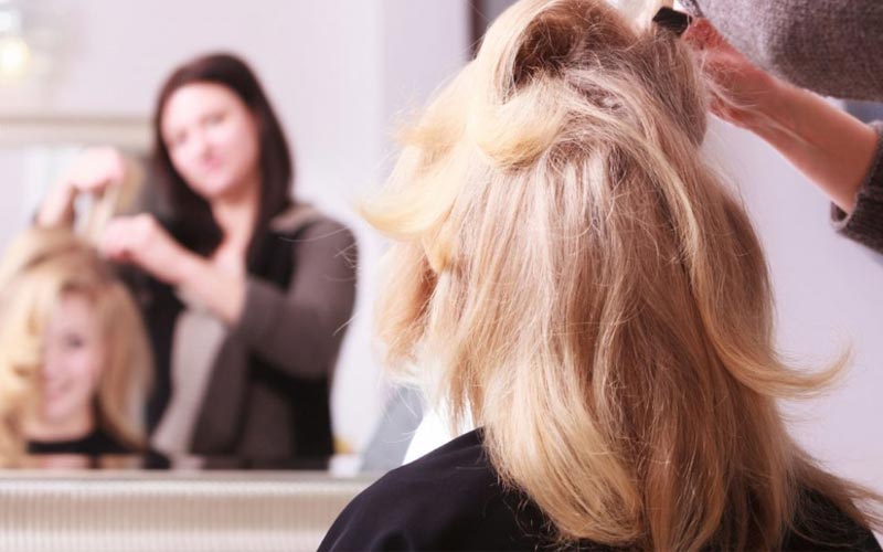 Femme blonde aux cheveux longs, se regardant dans le miroir, en train de se faire coiffer par une coiffeuse brune aux cheveux longs